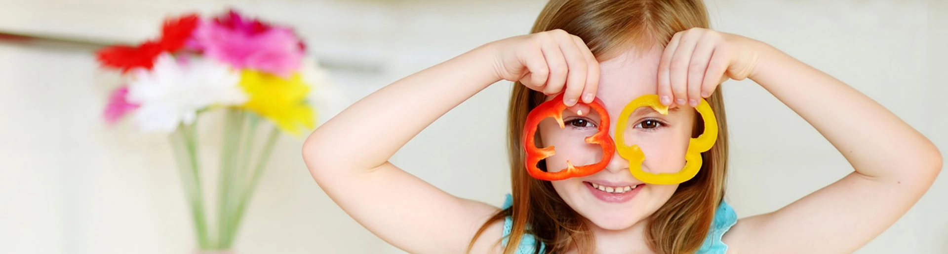 A girl playfully holds slices of red and yellow bell peppers over her eyes, smiling. A vase of colorful flowers is in the blurred background.