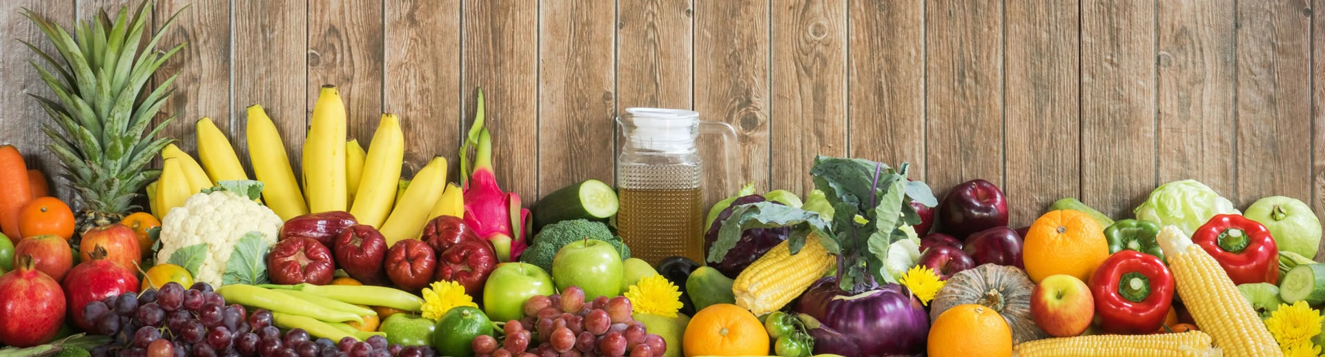 A variety of fruits and vegetables, including pineapples, bananas, grapes, and corn, are arranged in front of a wooden backdrop with a glass pitcher of juice in the center.