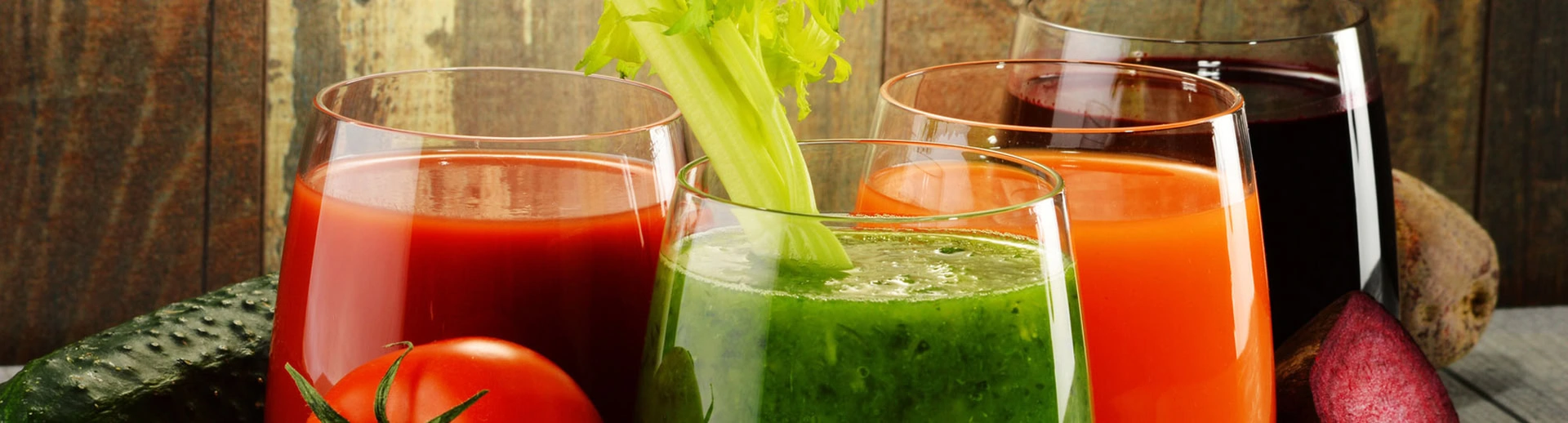 Four glasses of vegetable juices with garnishes, featuring green, orange, red, and purple colors, placed on a wooden surface with surrounding fresh vegetables.