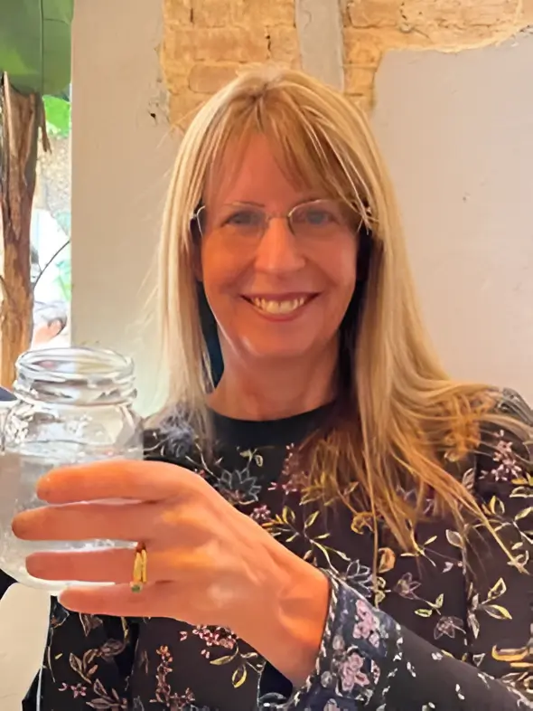 Woman with glasses, smiling and holding a jar with a clear liquid, wears a dark floral blouse. Background shows a light wall with some exposed brick.