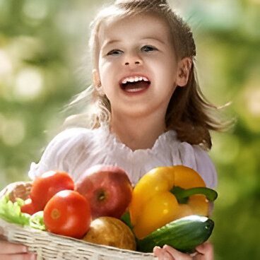 A smiling child holds a basket filled with vegetables and fruits, including a yellow pepper, tomatoes, apples, and a cucumber, outdoors.